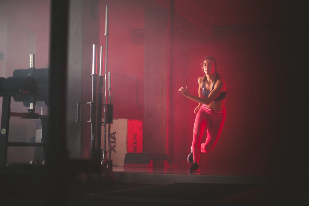 Eine Frau in Sportkleidung trainiert drinnen, joggt an Ort und Stelle unter dramatischer roter Beleuchtung in der Nähe von Fitnessgeräten und Gewichten - wie bei einer Sitzung mit einem Top Personal Trainer Hamburg.