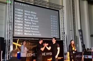 Zwei Männer schütteln sich auf der Bühne der HYROX-Veranstaltung in Hamburg die Hände. Die Namen und Zeiten der Teilnehmer - darunter auch Martins Erfolg - werden auf einer großen Leinwand angezeigt, während zwei weitere Personen in der Nähe zusehen.