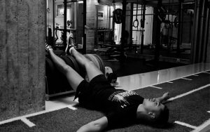 Eine Person in Sportkleidung liegt auf dem Rücken, die Beine sind an Fitnessgeräten angelehnt, die Arme sind ausgestreckt. Sie führt ein HIIT-Training in einem Fitnessstudio durch, in dem im Hintergrund Gewichte und Maschinen zu sehen sind.