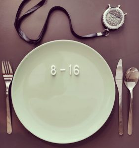 Ein Teller mit der Aufschrift "8 - 16", bestückt mit Besteck und einer Stoppuhr an einem schwarzen Band, stellt die Intermittent Fasting 16-8 Methode visuell dar und weist auf ihre Vor- und Nachteile hin.