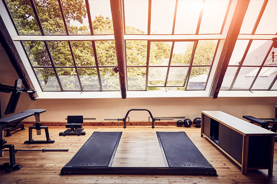 Ein modernes Fitnessstudio mit Holzböden, Trainingsbänken, zwei Laufbändern, Gewichten und großen, schrägen Fenstern, die das Sonnenlicht hereinlassen und das Grün draußen zeigen.