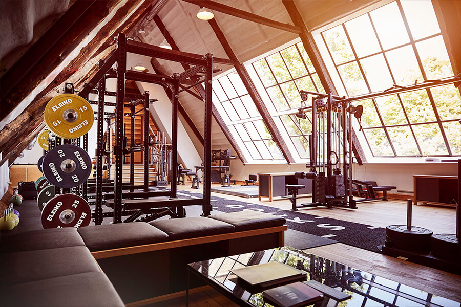Ein heller Fitnessraum mit großen schrägen Fenstern, Gewichthebern, Hanteln, Fitnessgeräten und Bänken, alles in einem großen Raum mit Holzböden und sichtbaren Dachbalken.