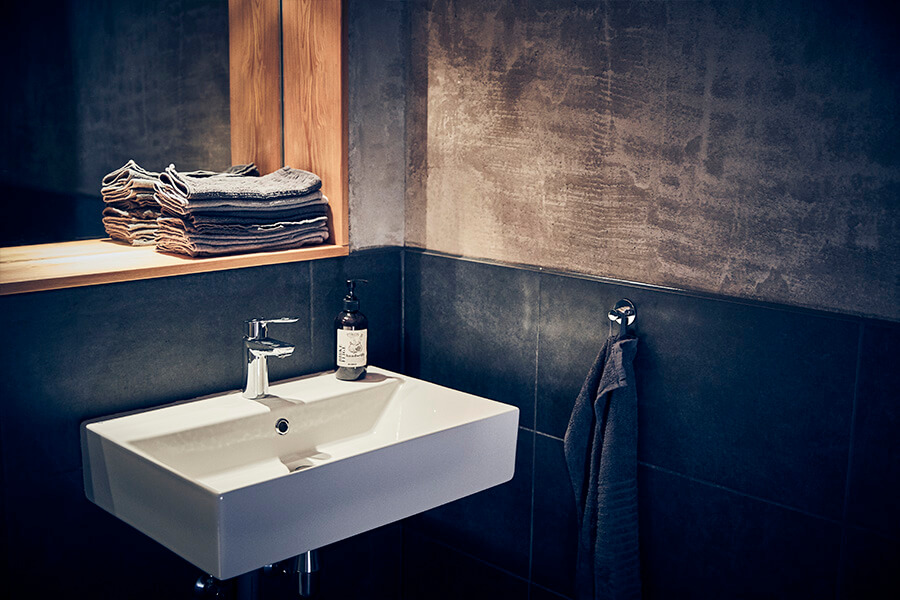 Ein modernes Badezimmer mit einem weißen, rechteckigen Waschbecken, einem verchromten Wasserhahn, einer Flasche Seife und einem dunklen Handtuch, das an der Wand hängt. Ein holzgerahmter Spiegel und ordentlich gefaltete Handtücher sind auf einem Regal über dem Waschbecken zu sehen.