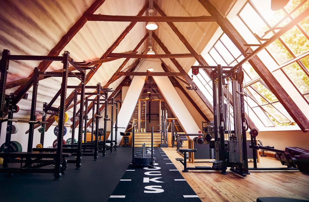 Ein modernes Fitnessstudio im Dachgeschoss mit freiliegenden Holzbalken, großen Fenstern, die das Sonnenlicht hereinlassen, verschiedenen Fitnessgeräten und einer Bahn in der Mitte, die zu einer Treppe führt.