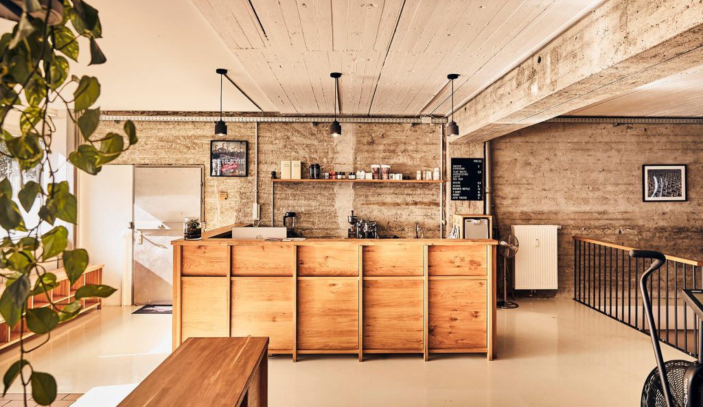 Ein modernes, minimalistisches Café mit Holztresen, Betonwänden und -decken, Topfpflanzen, Kaffeezubehör und gerahmter Kunst an den Wänden. Natürliches Licht erhellt den Raum.
