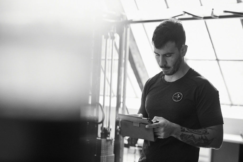 Ein Mann mit Tätowierungen und T-Shirt steht in einem Innenraum und blickt auf ein Tablet. Das Sonnenlicht strömt durch große Fenster hinter ihm und schafft eine helle, konzentrierte Atmosphäre.