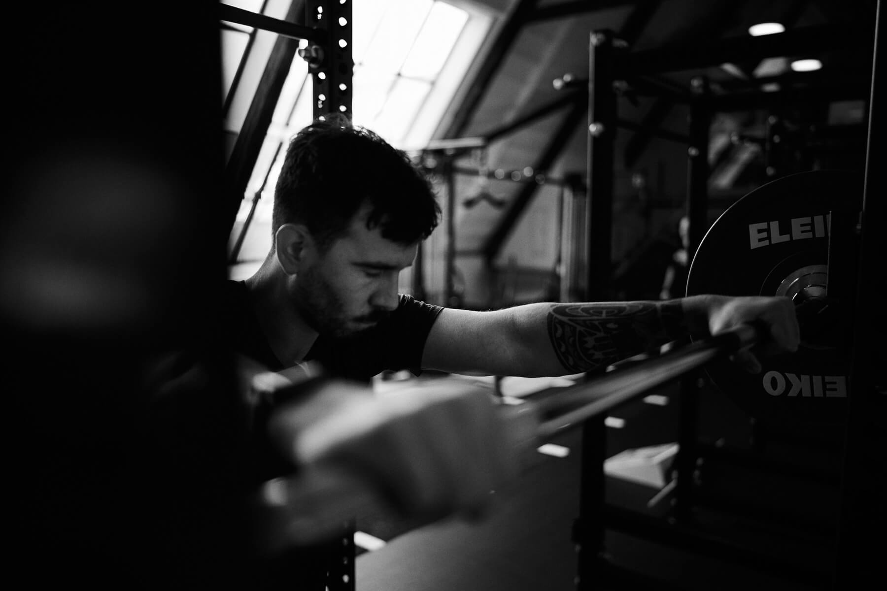 Ein Mann mit einer Tätowierung auf dem Arm hält eine Langhantel in einem Fitnessstudio. Er wirkt konzentriert und bereitet sich darauf vor, Gewichte zu heben. Das Bild ist in Schwarz-Weiß gehalten und hat eine weiche, stimmungsvolle Beleuchtung.