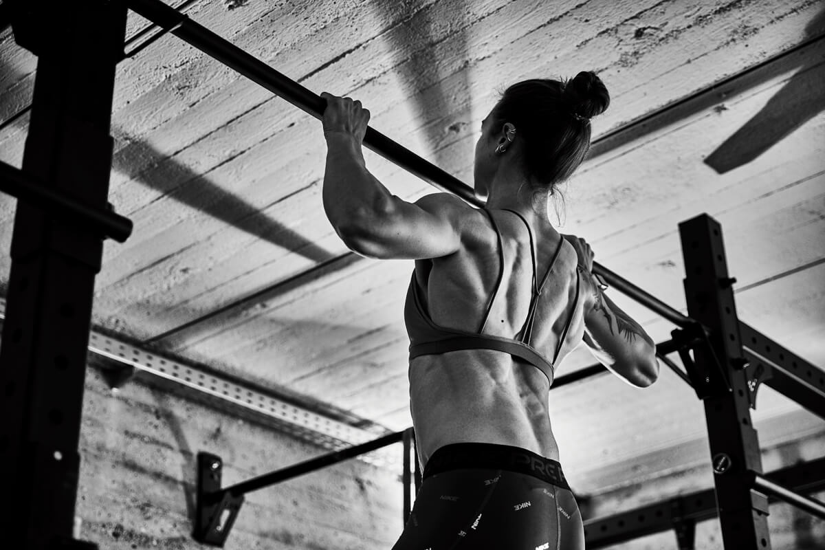 Eine Frau in Sportkleidung führt einen Klimmzug an einer Metallstange in einem Fitnessstudio durch und zeigt dabei einen muskulösen Rücken und definierte Arme. Das Bild ist in Schwarz-Weiß gehalten, wobei das Licht von oben einfällt.