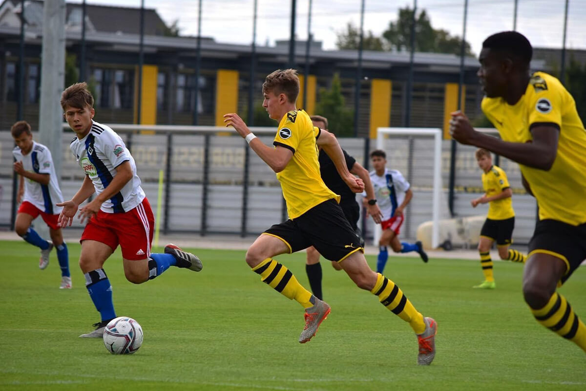 Mehrere junge Fußballspieler treten auf einem Rasenplatz gegeneinander an. Die Spieler in Gelb und Schwarz jagen dem Ball nach, während die Gegner in Weiß und Blau verteidigen. Im Hintergrund sind ein Stadion und ein Zaun zu sehen.