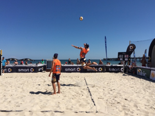 Zwei Männer spielen Beachvolleyball; einer springt, um den Ball zu schlagen, während der andere zusieht. Der Sandplatz liegt direkt am Meer, mit Zuschauern und einem Pier im Hintergrund unter einem klaren blauen Himmel.