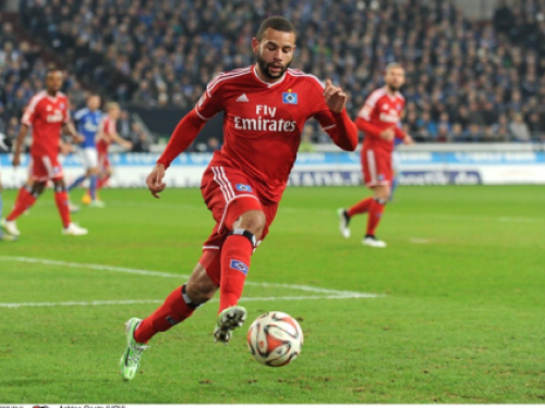 Ein Fußballspieler in einem roten Fly-Emirates-Trikot läuft während eines Spiels mit dem Ball auf einem grünen Feld, während die Zuschauer auf der Tribüne hinter ihm stehen.