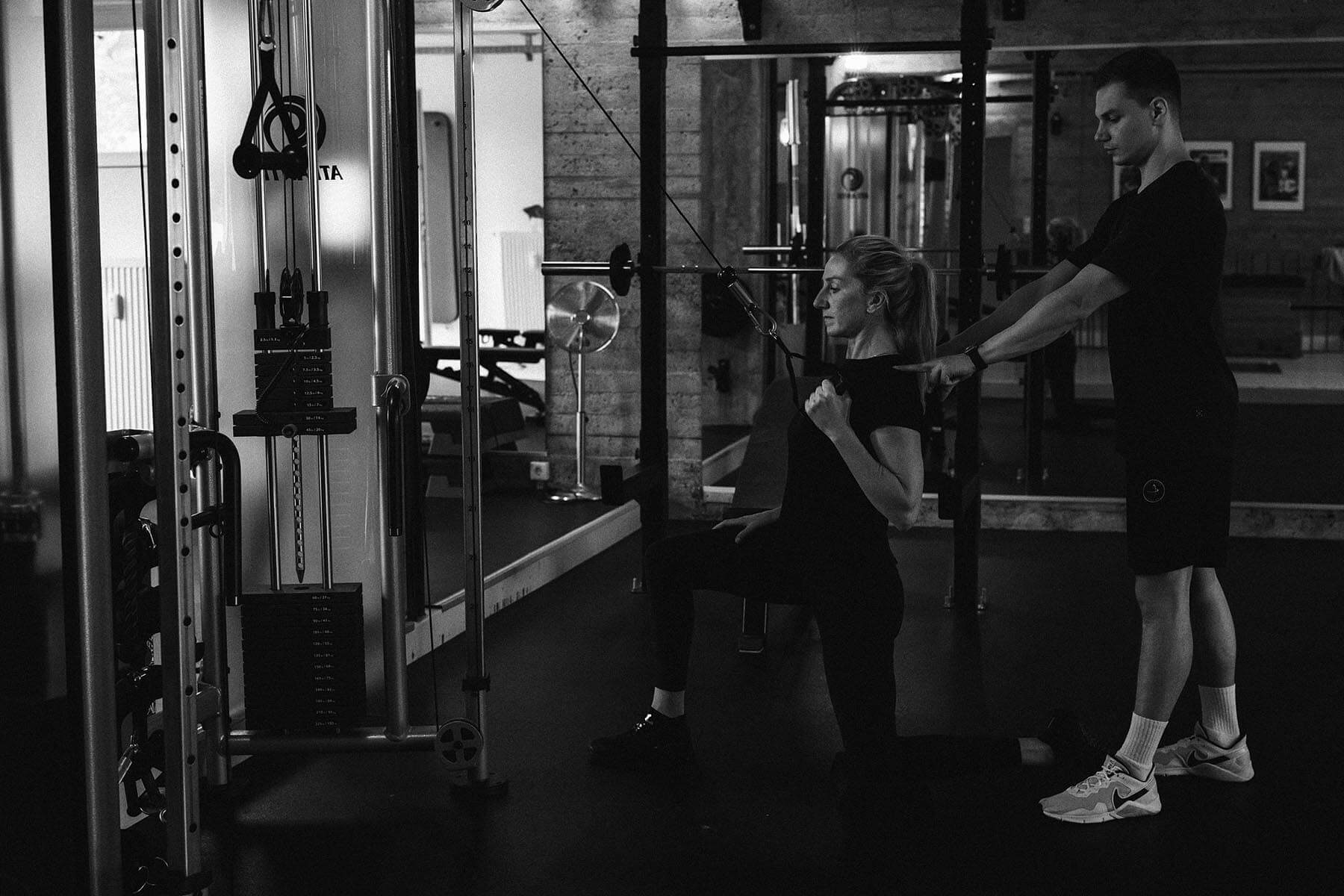 Eine Frau kniet beim Training an einem Kabelgerät und zieht den Griff an ihre Brust, während ein Mann hinter ihr steht und sie in einem Fitnessstudio unterstützt.
