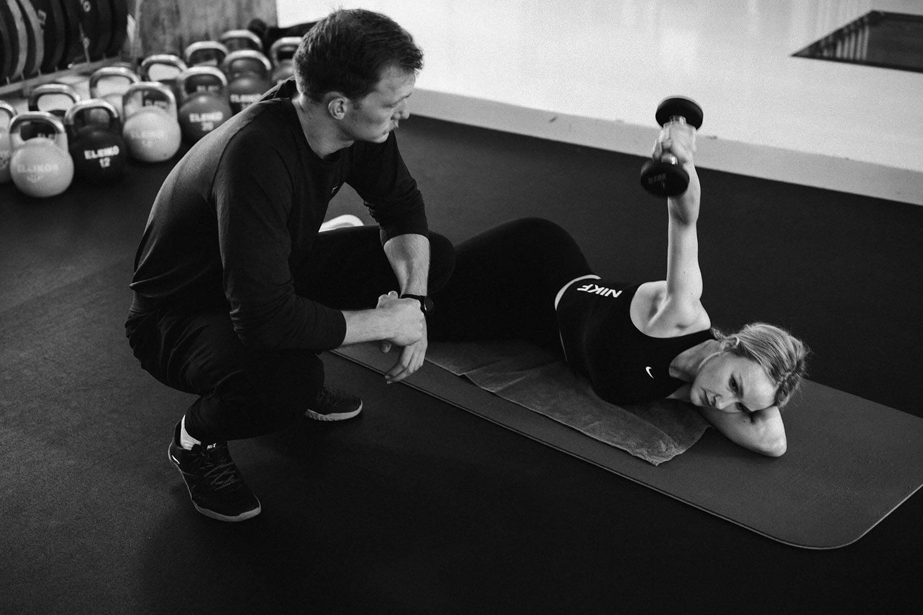Eine Frau, die auf einer Yogamatte liegt, hebt eine Hantel, während ein Trainer neben ihr kniet und sie in einem Fitnessstudio anleitet, in dem im Hintergrund Kettlebells aufgestellt sind.