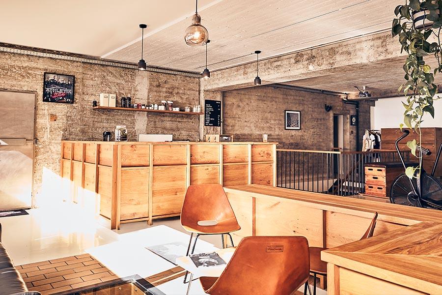 Ein modernes Café mit Holzmöbeln, braunen Stühlen, industriellen Betonwänden, Hängelampen und Topfpflanzen. Hinter dem hölzernen Tresen sind Regale mit Kaffeezubehör und eine Tafel mit der Speisekarte zu sehen. Durch die großen Fenster strömt Sonnenlicht.