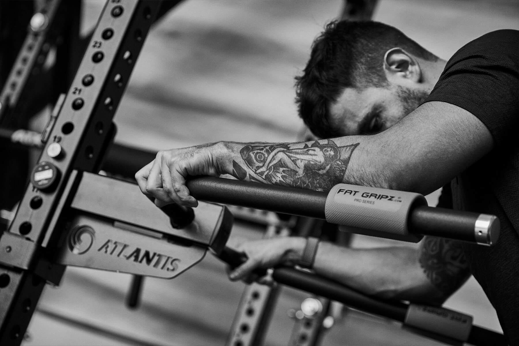 Ein Mann mit einer Tätowierung auf dem Unterarm stützt sich auf eine Fitness-Stange mit der Aufschrift FAT GRIPZ und ATLANTIS und wirkt erschöpft oder ruht sich während eines Trainings in einem Fitnessstudio aus. Das Bild ist in schwarz-weiß gehalten.