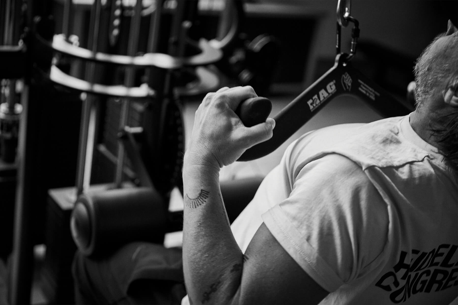 Eine Person in einem Fitnessstudio benutzt eine Kraftmaschine und hält den Griff mit einem tätowierten Unterarm fest. Das Bild ist in Schwarz-Weiß gehalten und konzentriert sich auf den Arm, die Schulter und einen Teil des Hemdes.