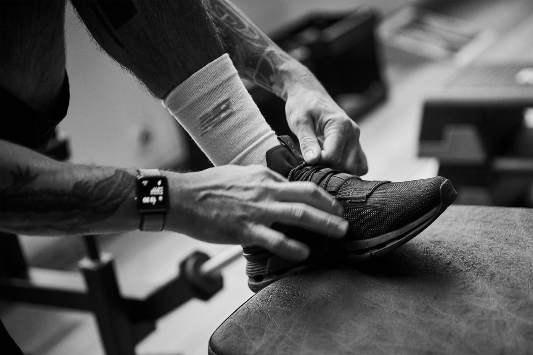 Eine Person, die eine Smartwatch trägt, bindet die Schnürsenkel eines schwarzen Sportschuhs, während sie ihren Fuß auf einer gepolsterten Bank abstützt, wobei im Hintergrund Fitnessgeräte zu sehen sind. Das Bild ist in schwarz-weiß gehalten.