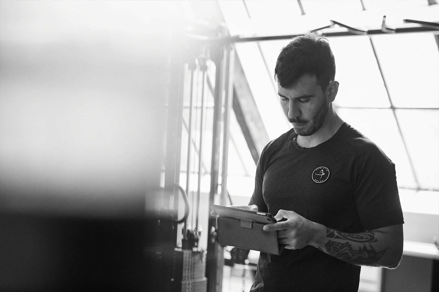 Ein Mann mit kurzen Haaren und Tätowierungen auf dem rechten Arm steht in einem Haus, schaut nach unten und benutzt ein Tablet. Er trägt ein dunkles T-Shirt mit einem kleinen Logo. Der Hintergrund ist leicht unscharf, was auf ein Fitnessstudio oder eine Fitnessumgebung hindeutet.