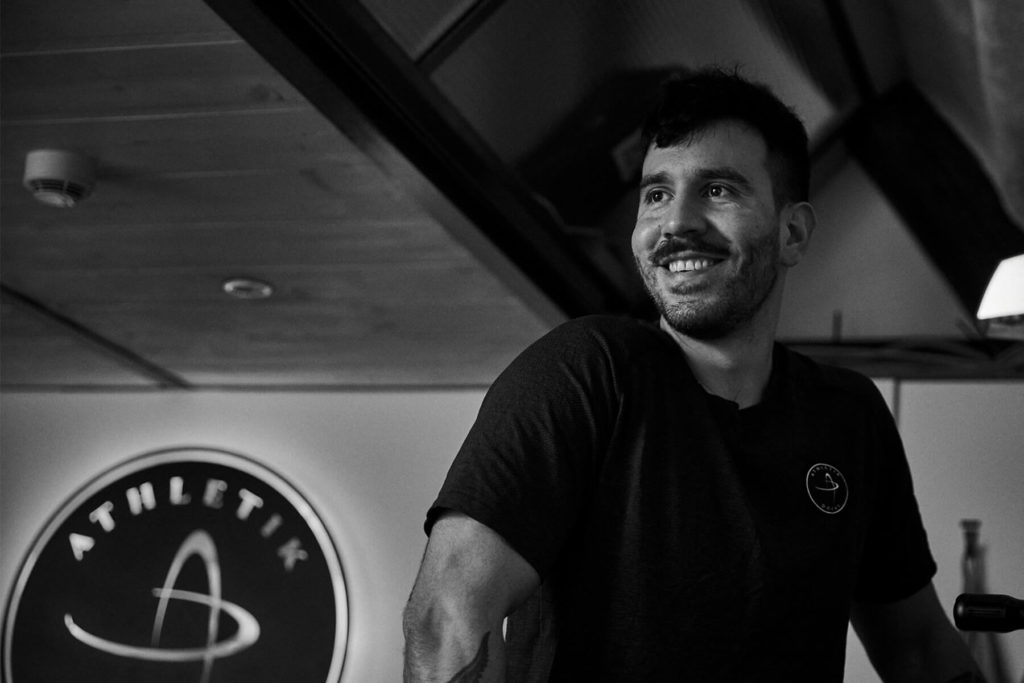 Ein Mann mit kurzen Haaren und Bart steht lächelnd in einem Innenraum und trägt ein dunkles T-Shirt. Im Hintergrund deutet ein rundes Schild mit der Aufschrift ATHLETIK auf seine Tätigkeit als Personal Trainer Hamburg hin. Das Foto ist in schwarz-weiß gehalten.