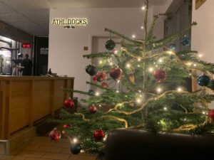 Ein geschmückter Weihnachtsbaum mit roten und grünen Ornamenten und Lichterketten steht im Vordergrund eines gemütlichen Innenraums, der perfekt für die Feiertage ist. Eine Holztheke und ein Schild mit der Aufschrift ATHL DOCKS zieren die Wand.
