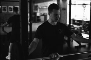 Ein Mann in einem Fitnessstudio, der ein dunkles T-Shirt trägt, steht neben einer Langhantel auf einer Hantelablage und bereitet sich auf das Heben vor. In dem Spiegel hinter ihm ist sein Spiegelbild zu sehen, das sich auf Lösungen für Diät-Stolpersteine konzentriert. Das Fitnessstudio ist schwach beleuchtet, die Geräte im Hintergrund sind unscharf.