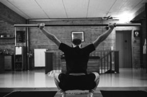 Eine Person in Sportkleidung führt eine Überkopfhocke mit einer Langhantel in einem Fitnessstudio aus, von hinten gesehen. Die Holzwände und die minimalistische Einrichtung schaffen eine konzentrierte Trainingsatmosphäre - ideal für Kalorien Tracking während intensiver Trainingseinheiten.