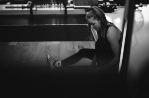 Eine Frau sitzt auf dem Boden eines Fitnessstudios und beugt sich vor, um ihren Sportschuh zu richten. Vorbereitet und konzentriert, mit ihrem zu einem Pferdeschwanz zurückgebundenen Haar, scheint sie entschlossen auf ihrer Abnehm-Reise. Das Bild ist in schwarz-weiß gehalten.