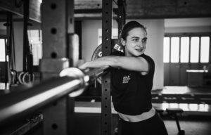 Eine Frau in Sportkleidung lehnt sich an eine Hantelablage in einem Fitnessstudio und lächelt, als wüsste sie: Auch wenn nicht dein Tag ist, gehört Leistungsfähigkeit einfach dazu. Das Schwarz-Weiß-Foto zeigt Fitnessgeräte im Hintergrund.