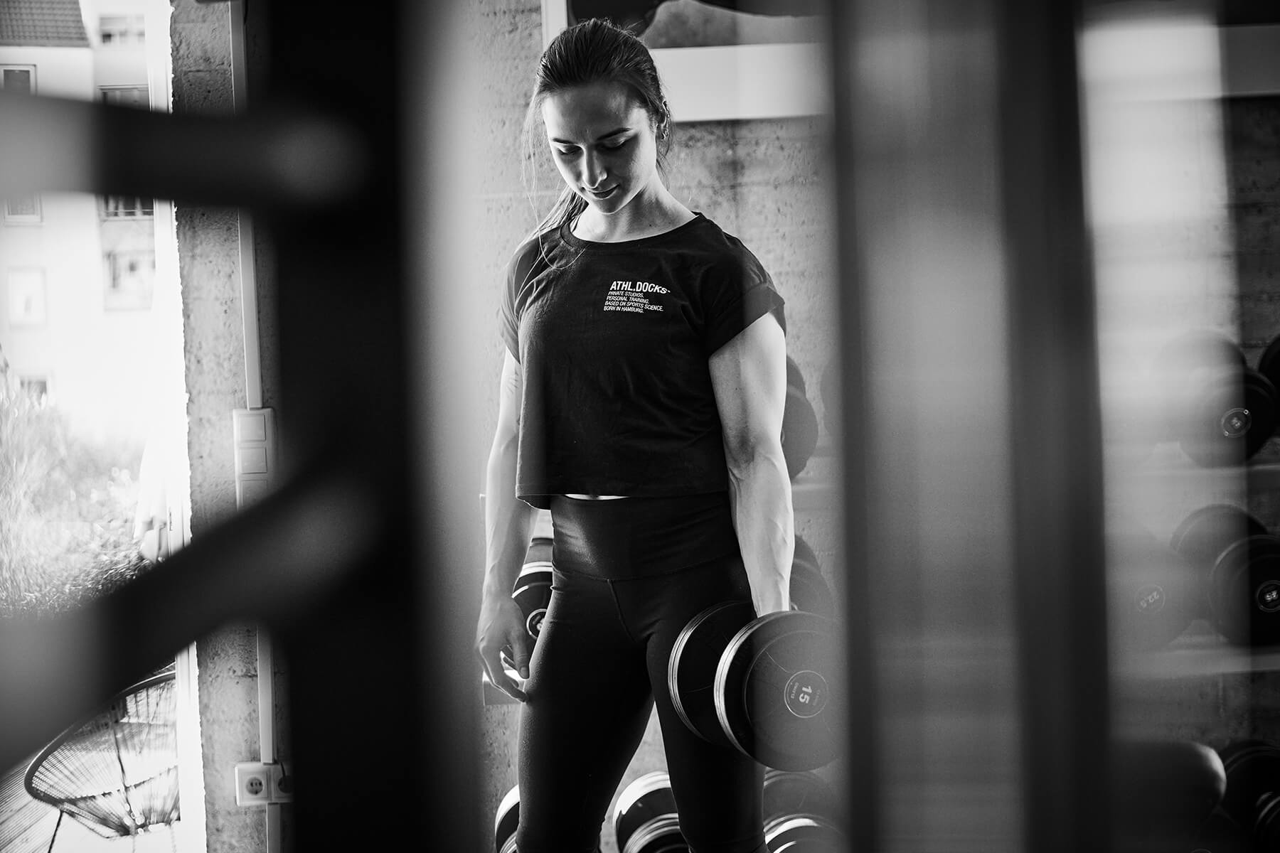 Eine Frau in Sportkleidung steht in einem Fitnessstudio und hält Hanteln, schaut nach unten und lächelt leicht. Das Bild ist schwarz-weiß, wobei die Fitnessgeräte im Vordergrund verschwimmen.