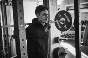 Eine Person mit Kapuzenpulli steht unter einer mit Eleiko 25 kg Hantelscheiben beladenen Langhantel und bereitet sich auf eine Kniebeuge in einem Fitnessstudio vor. Sie erinnert uns daran, dass Diätpausen und Training oft helfen, Plateaus beim Abnehmen zu überwinden und langfristig abnehmen zu können.