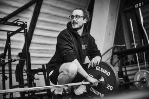 Eine Person mit Brille und Kapuzenpulli hockt neben einer belasteten Langhantel in einem Fitnessstudio und stützt sich lächelnd mit der Hand auf die Gewichte - eine Schwarz-Weiß-Szene, die die Freude am Trainingsfortschritt einfängt.