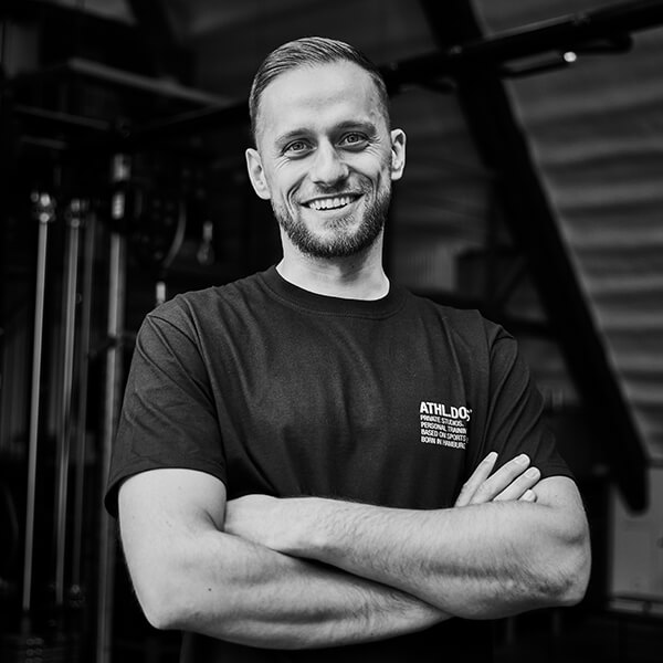 Ein Mann mit kurzen Haaren und einem Bart, der ein dunkles ATHLDG-T-Shirt trägt, steht mit verschränkten Armen in einer Turnhalle und lächelt in die Kamera.