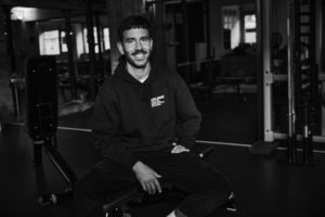 Ein Mann mit kurzen dunklen Haaren und einem Schnurrbart, bekleidet mit einem Kapuzenpulli und einer Hose, sitzt auf einer Bank und lächelt in einem Fitnessstudio. Das Schwarz-Weiß-Foto fängt die schwach beleuchtete Umgebung ein und hebt die Fitnessgeräte und seine Leidenschaft für Krafttraining hervor.