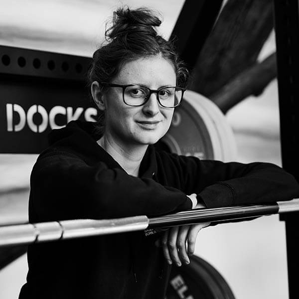 Eine Frau mit Brille und Duttfrisur stützt sich lässig auf eine Langhantel in einem Fitnessstudio, im Hintergrund sind Hantelscheiben und Fitnessgeräte zu sehen. Das Foto ist in schwarz-weiß gehalten.