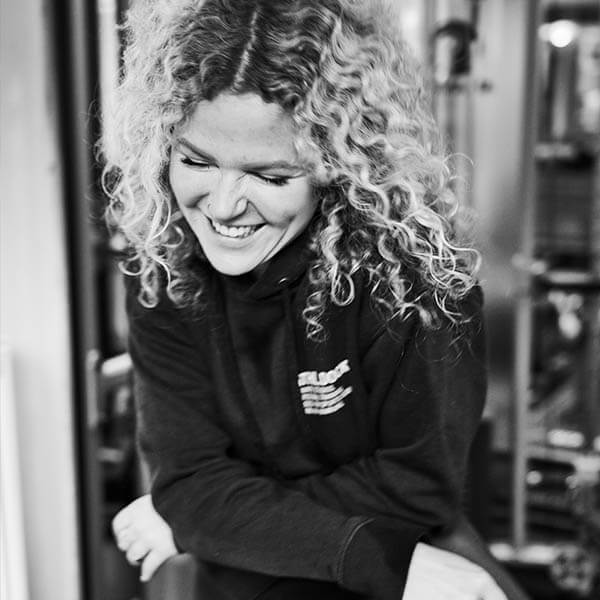 Schwarz-Weiß-Foto einer Frau mit lockigem Haar, die einen Kapuzenpulli trägt, lächelt und nach unten schaut, während sie ihre Arme verschränkt, in einem scheinbar geschlossenen Raum.