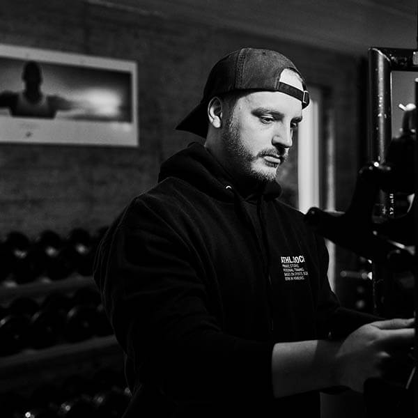 Ein Mann mit Kapuzenpulli und umgekehrter Mütze steht in einem Fitnessstudio und stellt Geräte ein. Im Hintergrund sind eine Ablage mit Hanteln und ein gerahmtes Foto an der Wand zu sehen. Das Bild ist in schwarz-weiß gehalten.