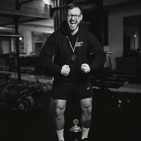 Ein Mann mit Brille und Medaille steht in einer Turnhalle, lächelt und reckt die Arme in die Höhe. Er ist mit einem Kapuzenpulli und Shorts bekleidet und hat eine Trophäe vor sich auf dem Boden stehen. Im Hintergrund sind Fitnessgeräte zu sehen.