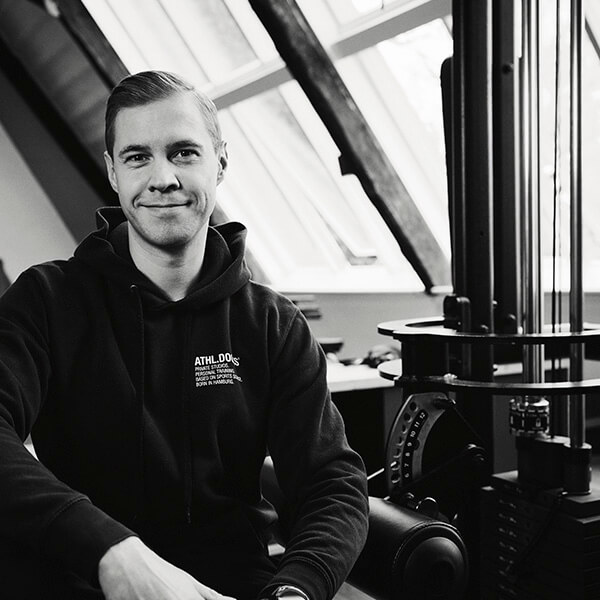 Ein Mann im Kapuzenpulli sitzt lächelnd in einem sonnendurchfluteten Raum mit schrägen Fenstern. Neben ihm steht ein Fitnessstudio oder ein Trainingsgerät. Das Bild ist in schwarz-weiß gehalten.