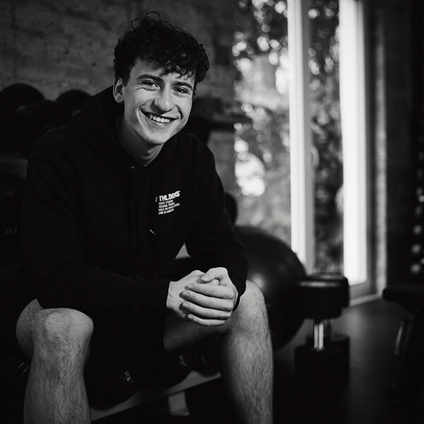 Ein junger Mann mit lockigem Haar, der einen Kapuzenpulli und kurze Hosen trägt, sitzt auf einer Bank in einem Fitnessstudio und lächelt in die Kamera. Im Hintergrund sind Hanteln und große Fenster zu sehen. Das Bild ist in schwarz-weiß gehalten.