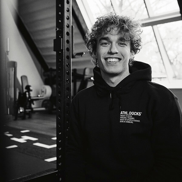 Ein junger Mann mit lockigem Haar lächelt in die Kamera, während er in einer Turnhalle sitzt. Er trägt einen Kapuzenpullover mit der Aufschrift "ATHL. DOCKS*" und im Hintergrund sind Trainingsgeräte zu sehen. Das Bild ist in schwarz-weiß gehalten.