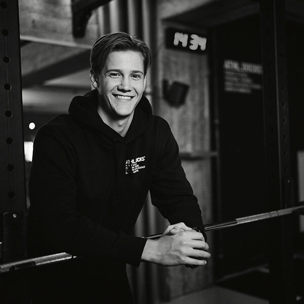 Ein junger Mann in einem schwarzen Kapuzenpulli lächelt, während er hinter einer Hantel in einem Fitnessstudio steht. Im Hintergrund sind Fitnessgeräte und eine Digitaluhr zu sehen, die die Zeit 14:34 anzeigt. Das Foto ist in schwarz-weiß gehalten.