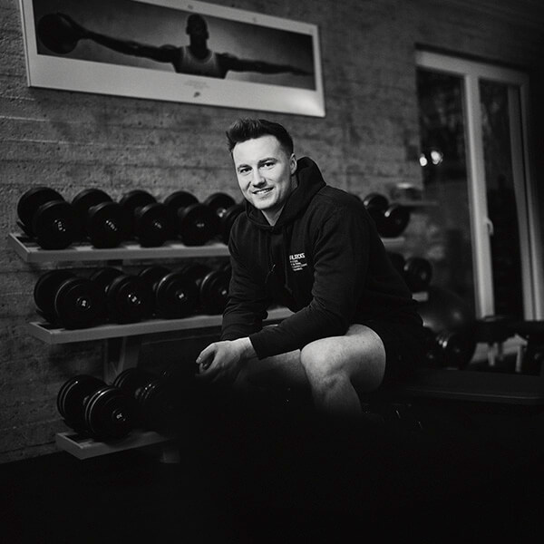 Ein Mann im Kapuzenpulli sitzt auf einer Bank in einem Fitnessstudio und lächelt in die Kamera. Hinter ihm stehen Hantelständer und an der Wand hängt ein gerahmtes Foto eines Gewichthebers. Das Bild ist schwarz-weiß.