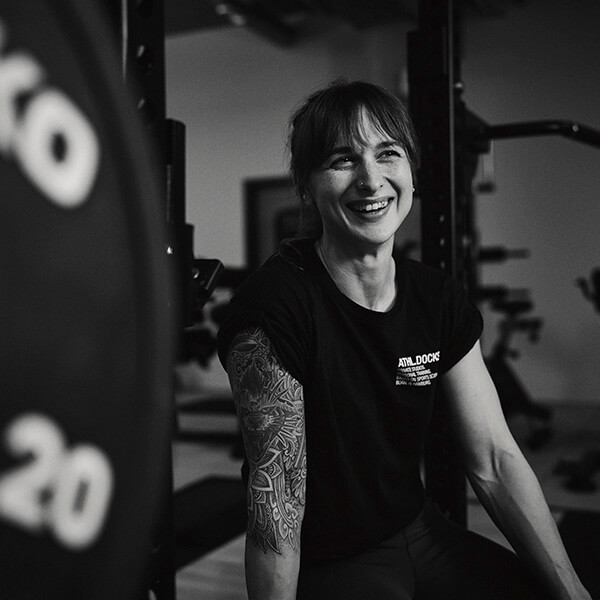 Eine Frau mit einem tätowierten Arm sitzt in einem Fitnessstudio und lächelt, sie trägt ein dunkles T-Shirt. Im Hintergrund sind Gewichthebegeräte und Gewichte zu sehen. Das Bild ist in schwarz-weiß gehalten.