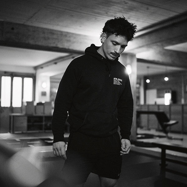 Ein Mann in Kapuzenpulli und Shorts steht in einem Fitnessstudio und schaut mit konzentriertem Blick nach unten. Das Schwarz-Weiß-Foto hebt den modernen, industriell anmutenden Trainingsraum um ihn herum hervor.