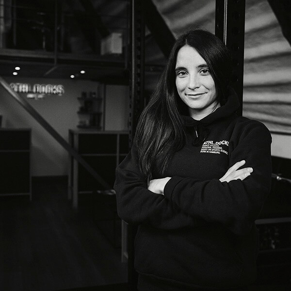 Eine Frau mit langen dunklen Haaren steht mit verschränkten Armen in einem Raum und trägt einen dunklen Kapuzenpullover. Sie lächelt leicht, und im Hintergrund ist ein Büro im Industriestil mit Metallträgern und Regalen zu sehen.