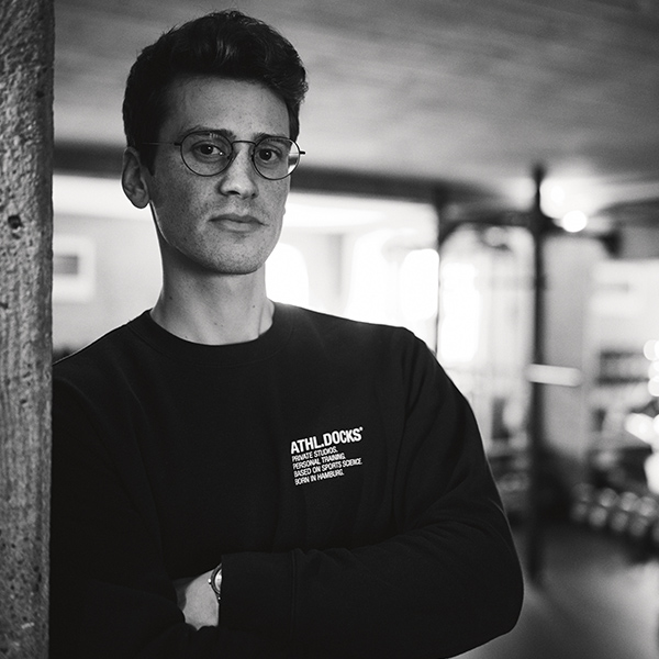 Ein junger Mann mit kurzen Haaren und Brille steht mit verschränkten Armen in einem Innenraum des Studio Sternschanze und trägt ein dunkles Sweatshirt mit der Aufschrift ATHLDOCKS. Der Hintergrund ist sanft verschwommen und wird von natürlichem Licht beleuchtet.