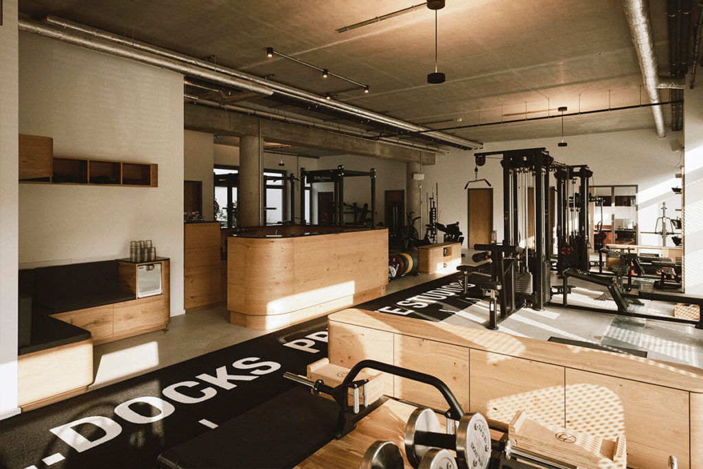 Ein modernes Fitnessstudio mit großen Fenstern, Holzakzenten, schwarz-weißen Fitnessgeräten, freien Gewichten, Maschinen, Bänken und einer zentralen Rezeption in einem geräumigen, hellen, offenen Bereich.