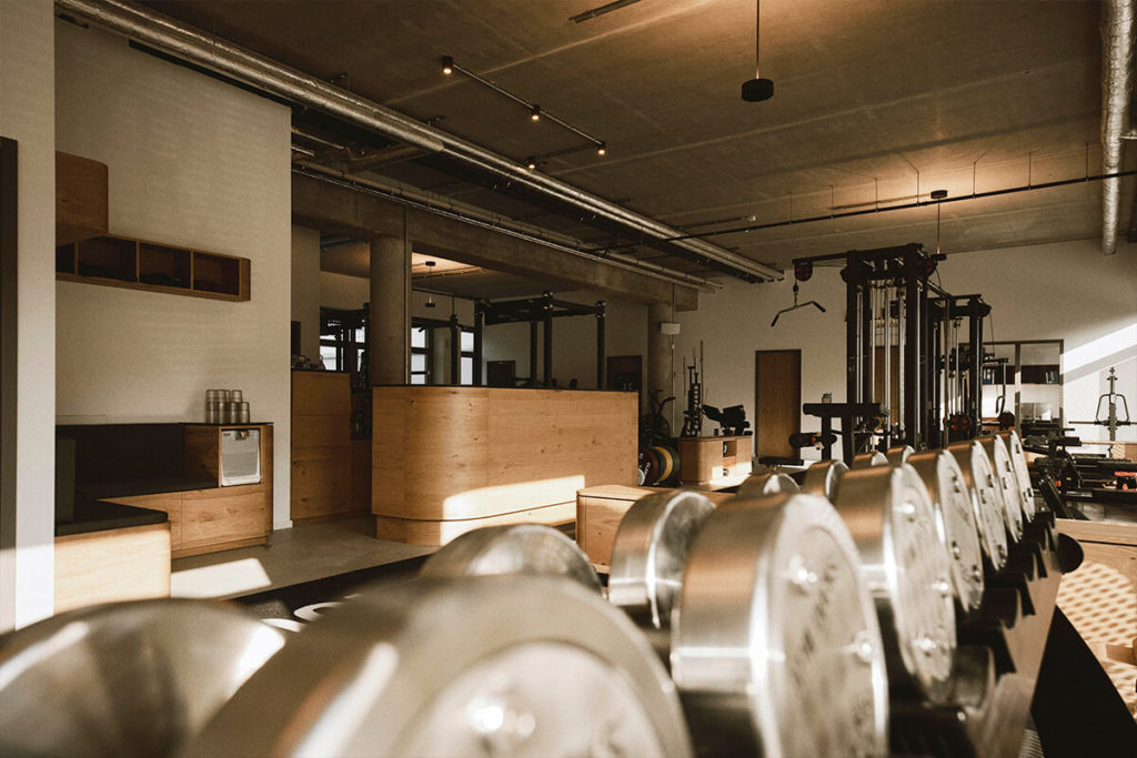 Modernes Fitnessstudio mit Reihen glänzender Hantelscheiben im Vordergrund, Geräten aus Holz und Metall, großen Fenstern, freiliegenden Decken und Beleuchtung im Industriestil. Warmes Sonnenlicht durchflutet den Raum.