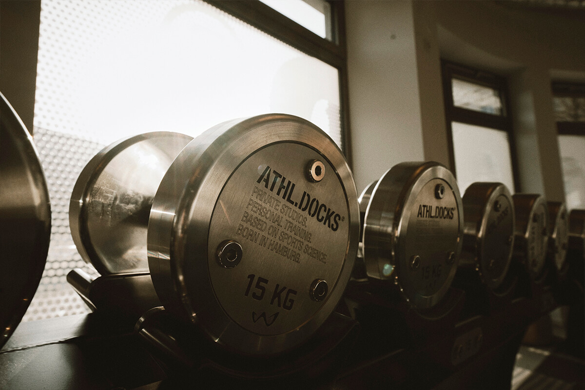 Eine Reihe metallisch glänzender Hanteln mit der Aufschrift ATHL.DOCKS und einem Gewicht von 15 kg ist auf einem Gestell an einem sonnenbeschienenen Fenster in einem Fitnessstudio angeordnet.