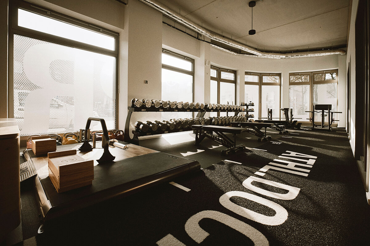 Ein modernes Fitnessstudio mit großen Fenstern, freien Gewichten, Bänken, Kettlebells und Boxen, das in natürliches Licht getaucht ist. Der Boden ist mit fetter weißer Schrift markiert und der Raum ist minimalistisch und sauber gestaltet.